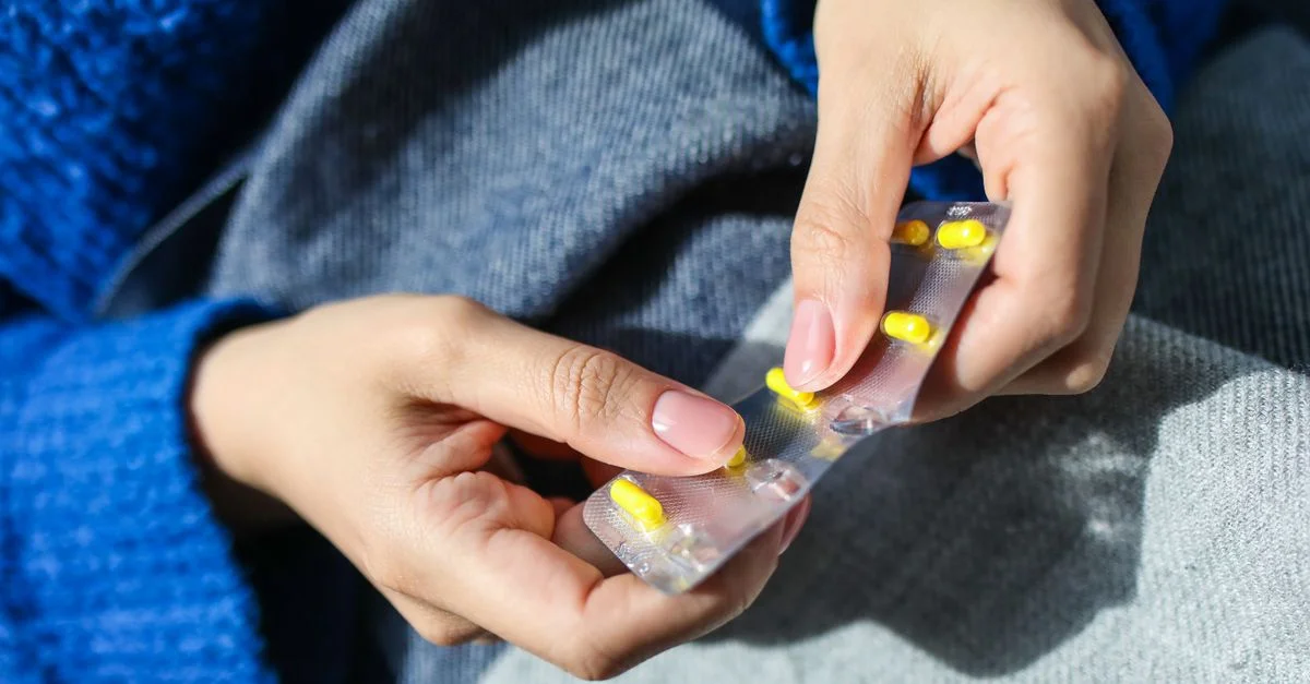 Close-up de las manos de una mujer sosteniendo un blister con pastillas amarillas, en interiores.