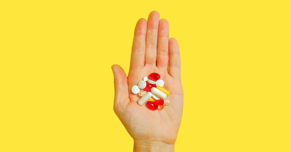 Primer plano de una mano adulta sosteniendo diferentes tipos de pastillas contra un fondo amarillo brillante.