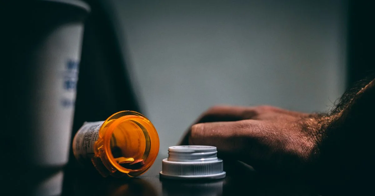 Escena iluminada de manera melancólica de una botella de receta abierta y una mano sobre una mesa, que muestra temas de atención médica.