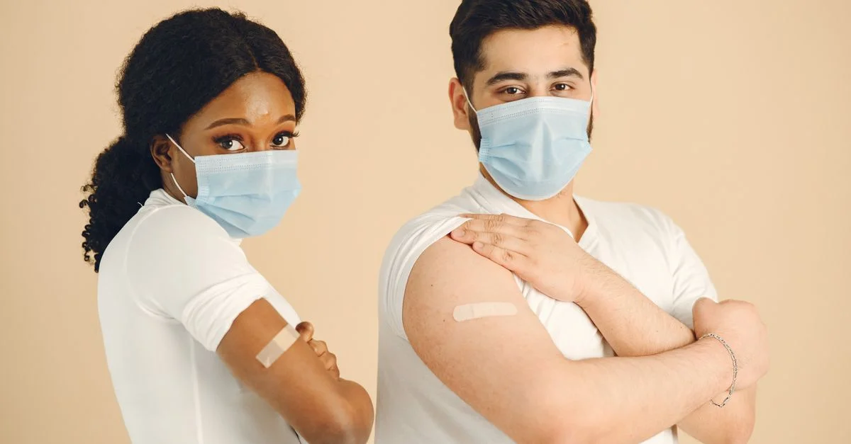Dos adultos con mascarillas muestran orgullosamente los vendajes de vacunación en sus brazos.