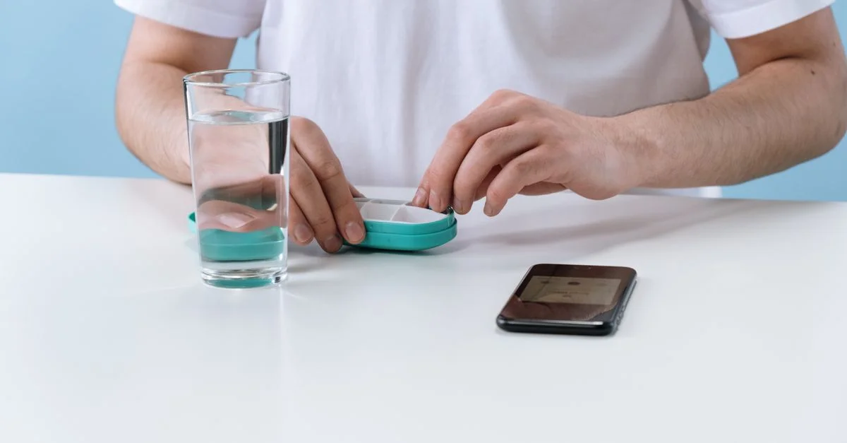 Una persona organizando pastillas con un organizador semanal, junto a un vaso de agua y un teléfono inteligente.