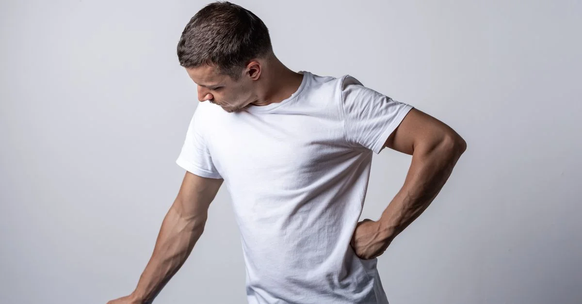 Un hombre con camiseta blanca se apoya en una silla azul frente a un fondo neutro.