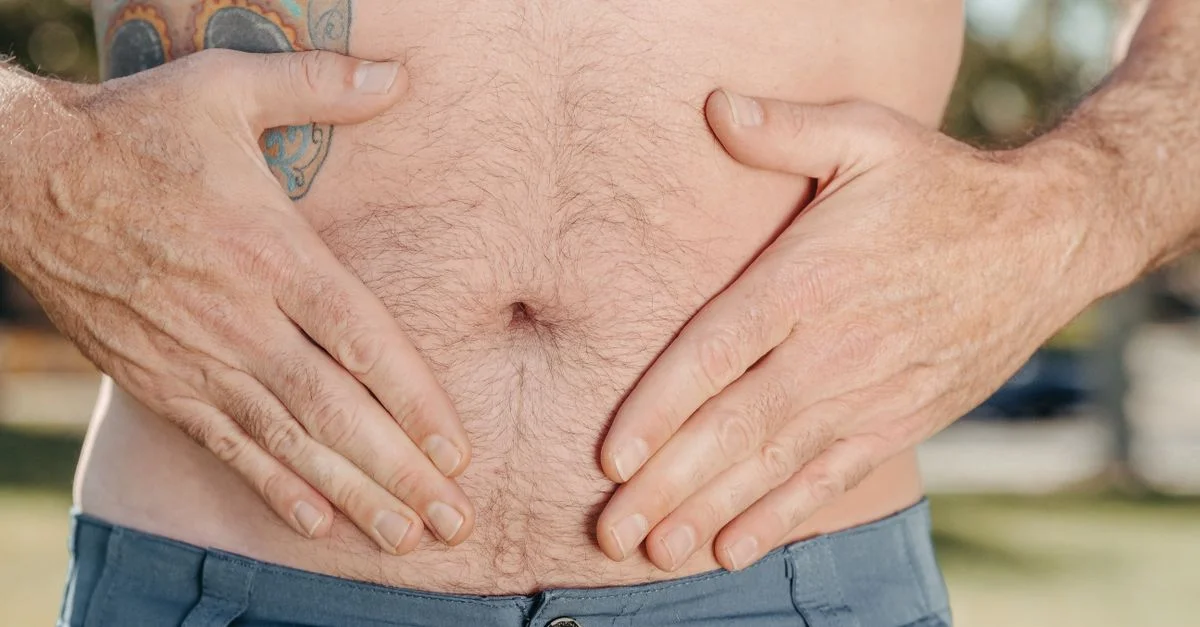 Una imagen en primer plano del estómago de un hombre con tatuajes, mostrando manos a cada lado, simbolizando salud o malestar.