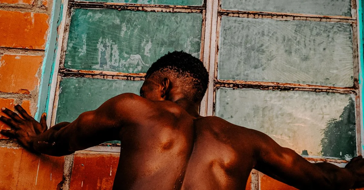 Un hombre sin camiseta se apoya en una ventana desde atrás, mostrando su espalda musculosa.