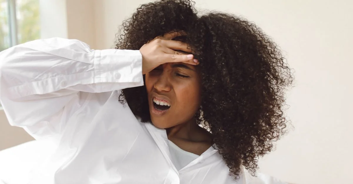 Una joven con cabello rizado despertando con dolor de cabeza. Lleva una camisa blanca y parece estar experimentando incomodidad.