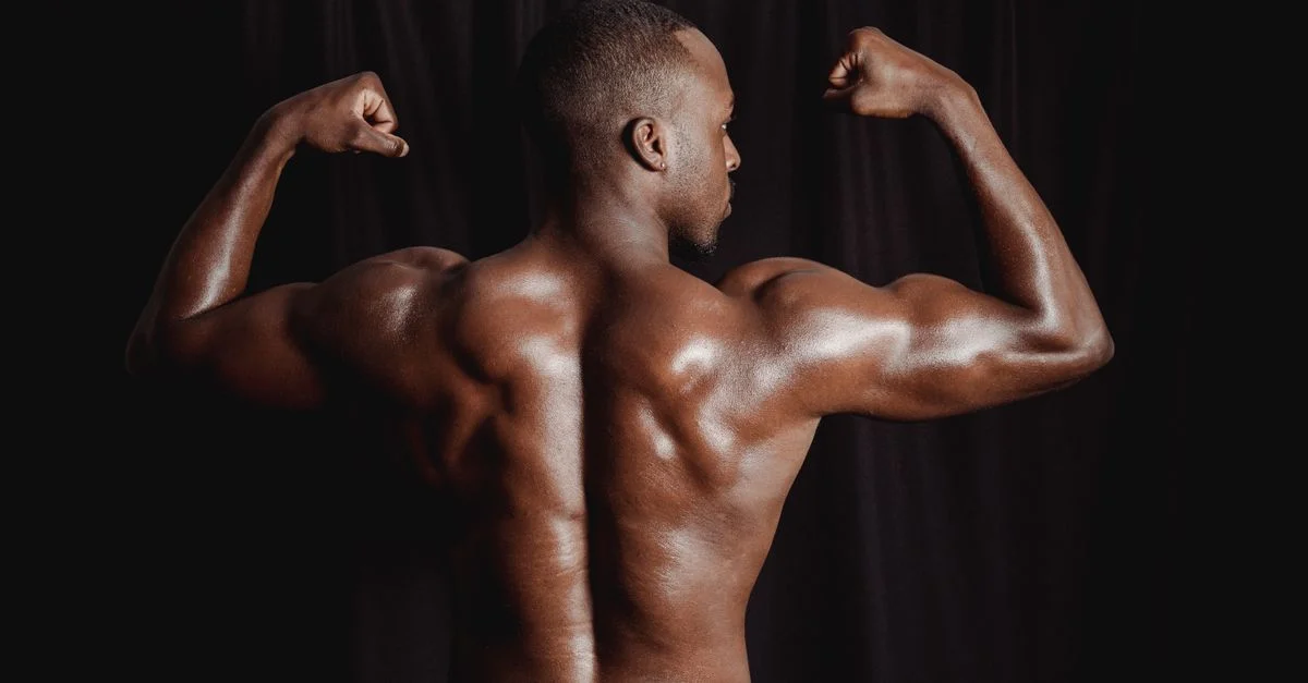 Hombre negro musculoso adulto flexionando los músculos de la espalda contra un fondo oscuro, mostrando fuerza y estado físico.