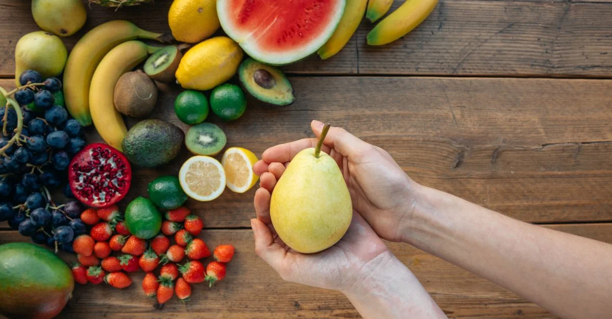 Manos sosteniendo una pera rodeada de una colorida variedad de frutas sobre una superficie de madera.