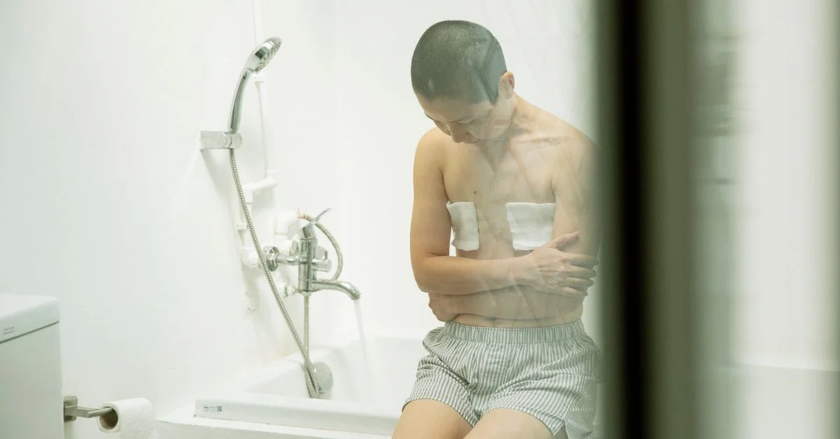 Mujer asiática sentada pensativa en una bañera con vendajes en el pecho, evocando una sensación de introspección y recuperación.